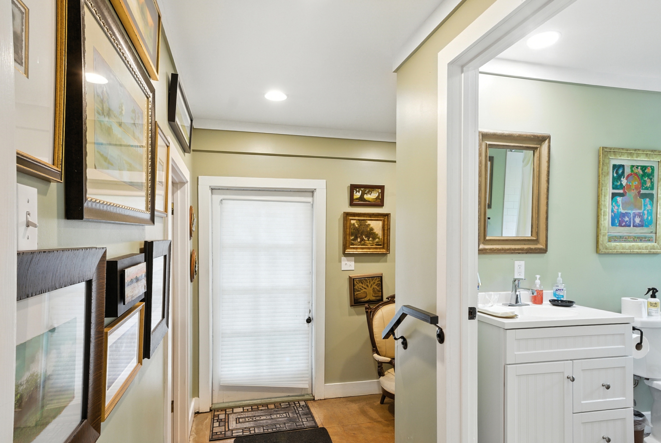 Sage green hallway featuring a gallery wall of framed artwork, hardwood floors, and an open doorway leading to a bathroom with a white vanity.