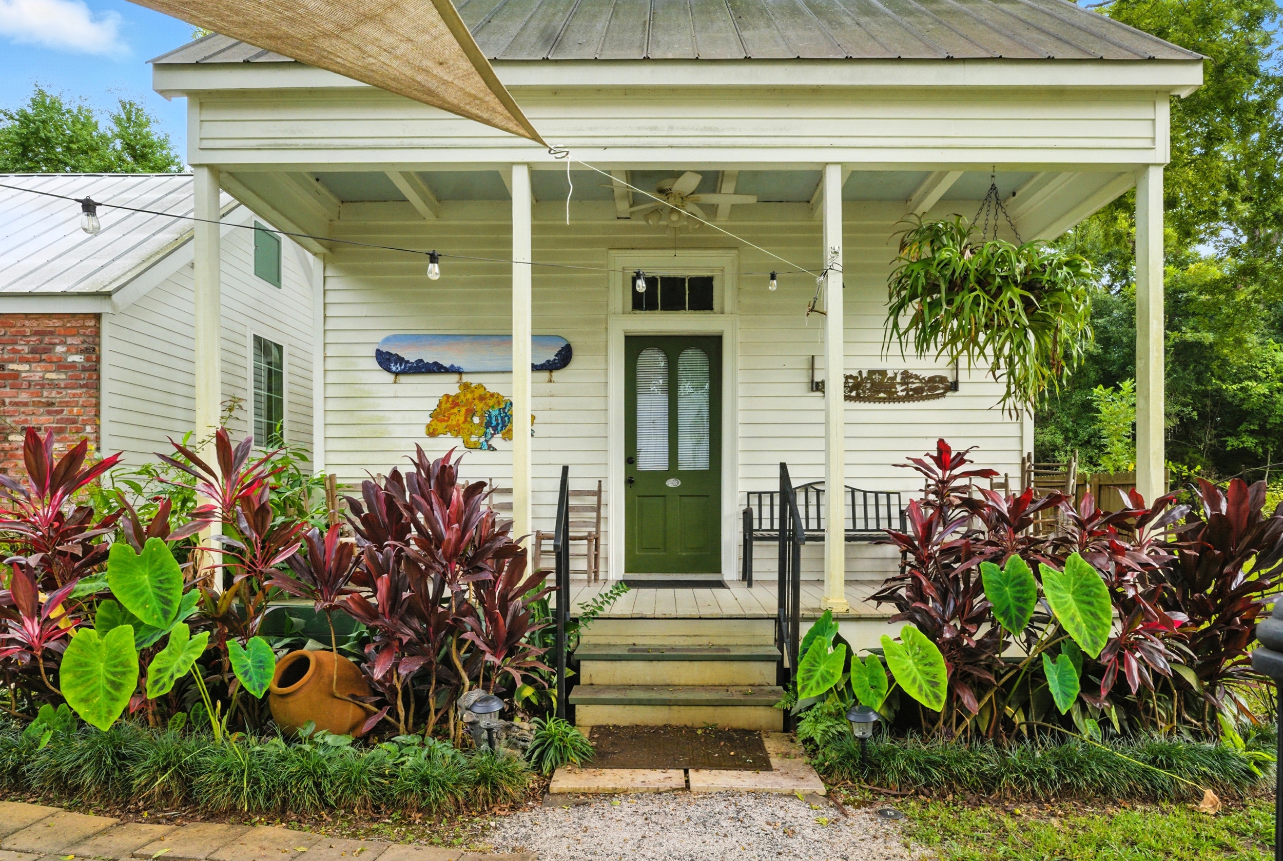 Inviting front porch of a white cottage featuring a green door, hanging ferns, and lush tropical landscaping.