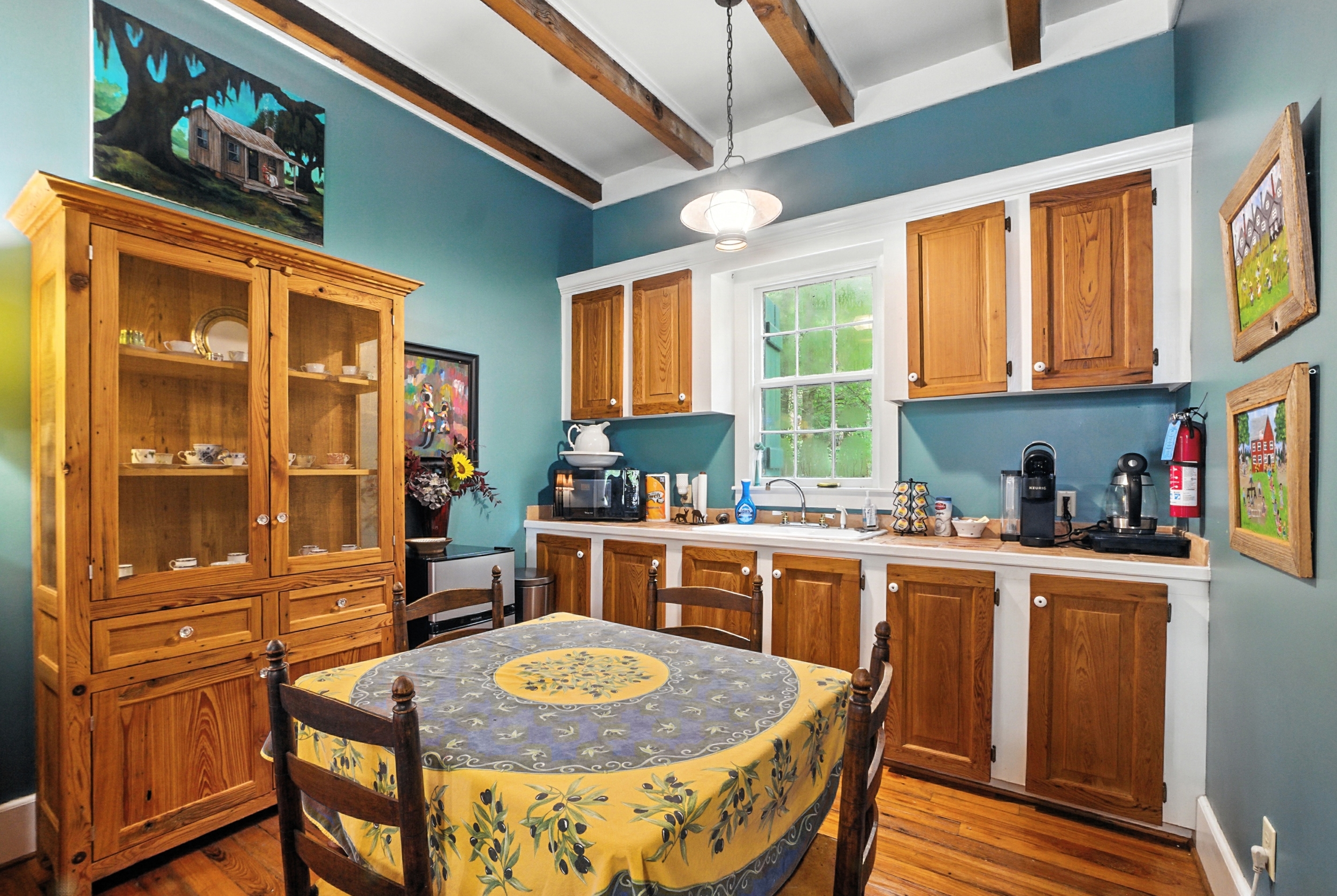 Cozy kitchenette with teal walls and exposed beams, featuring wooden cabinetry, a small dining table with a patterned tablecloth, and a large wooden hutch.