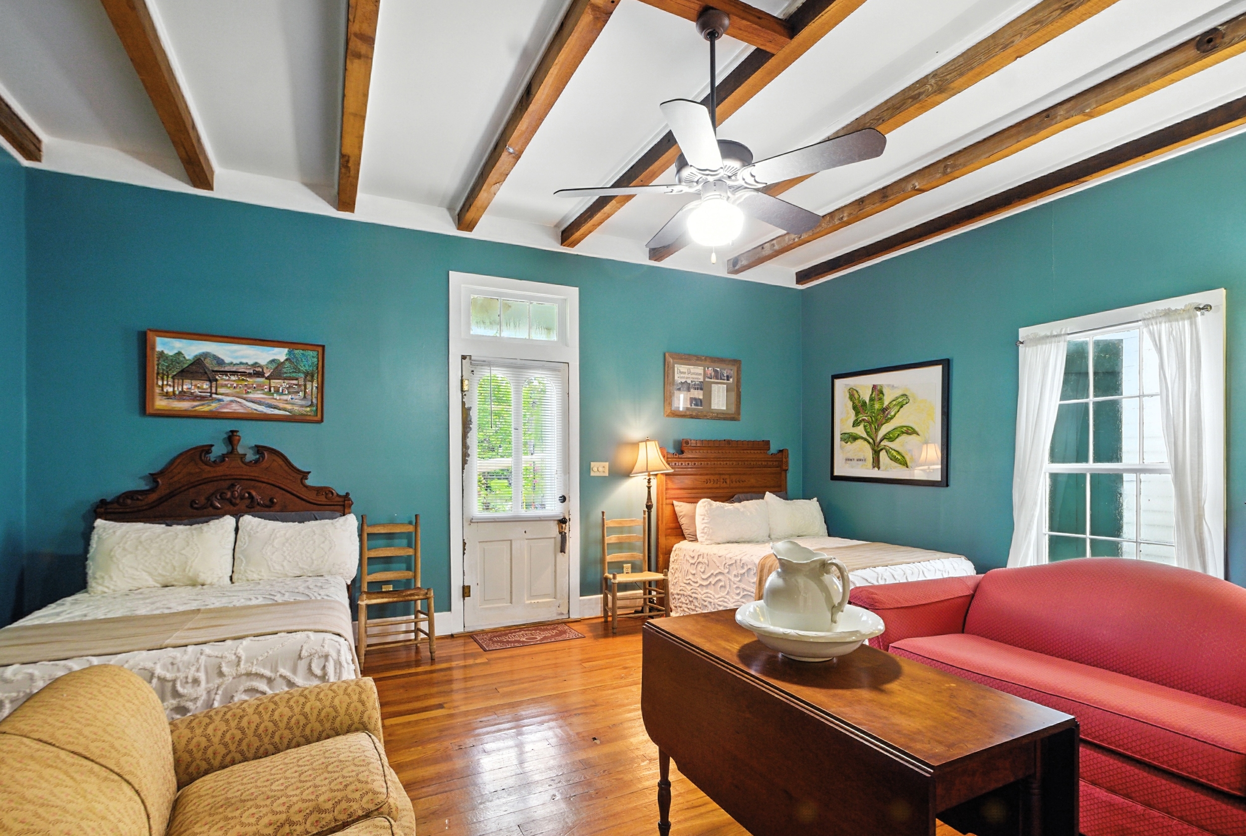 Spacious teal bedroom featuring two beds with wooden headboards, exposed ceiling beams, hardwood floors, a pink sofa, and a wooden drop-leaf table.