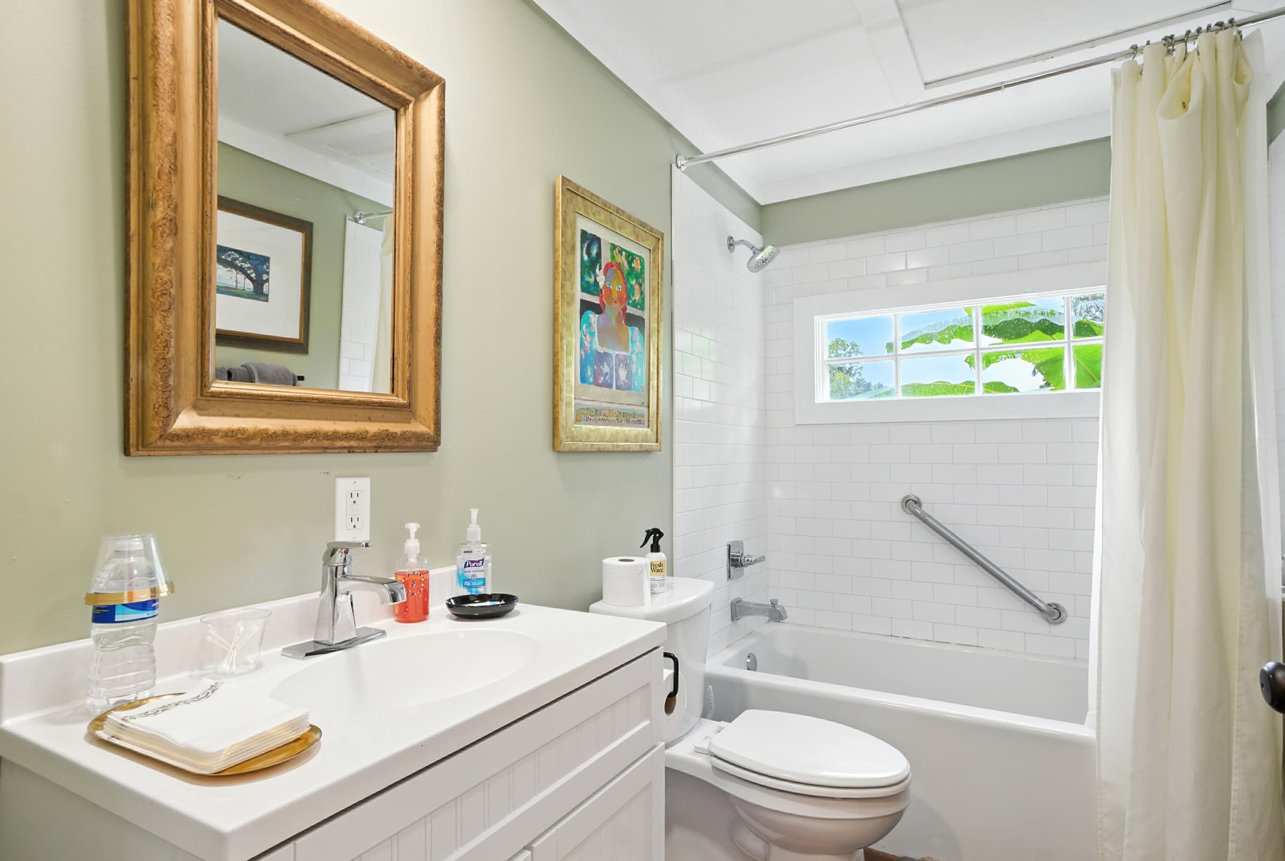 Bright bathroom featuring sage green walls, a white vanity with a gold-framed mirror, and a tiled shower/tub combo with a window.