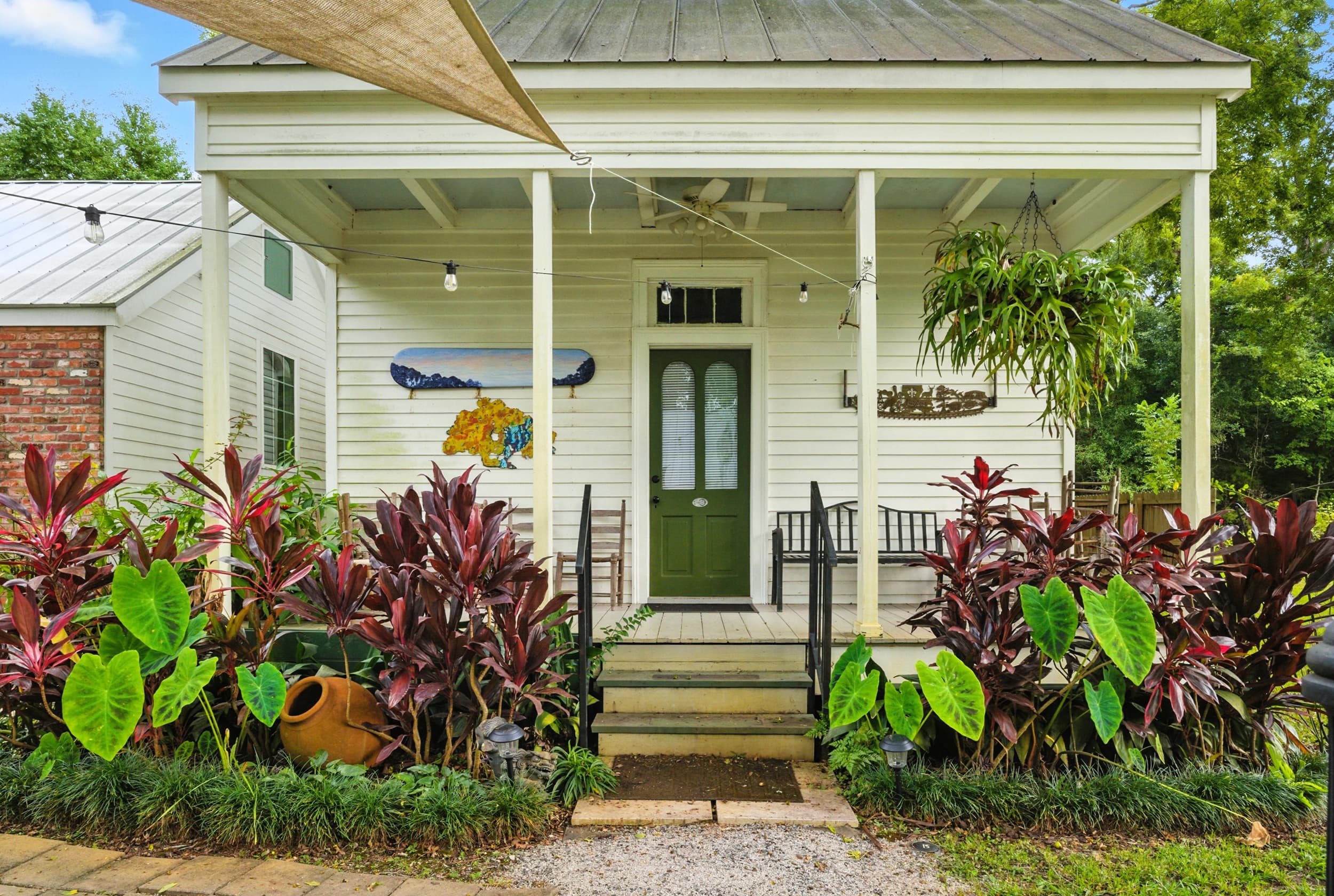 Inviting front porch of a white cottage featuring a green door, hanging ferns, and lush tropical landscaping.
