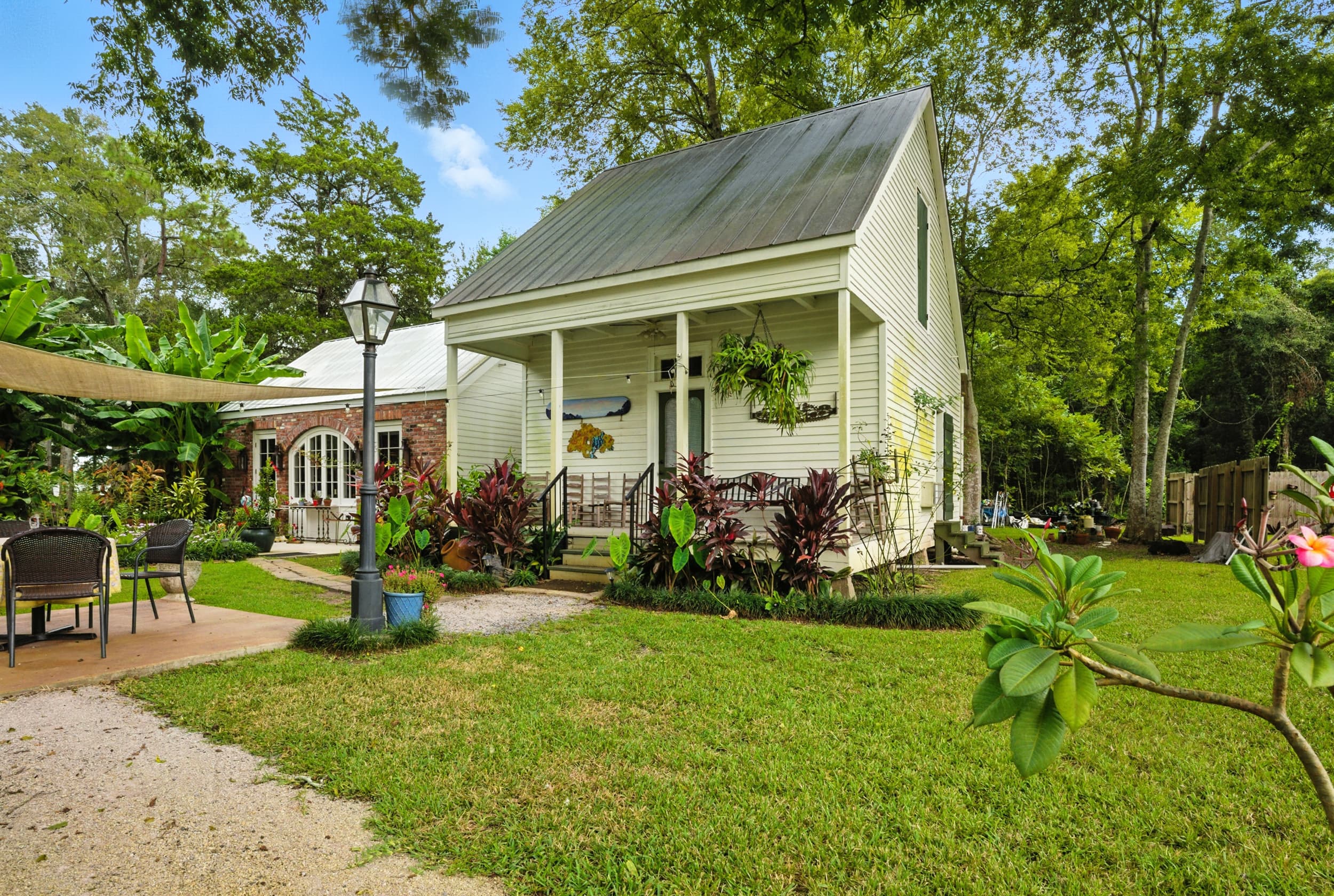 Exterior view of a charming white cottage featuring a metal roof, inviting front porch, and a spacious garden lawn with a patio area.
