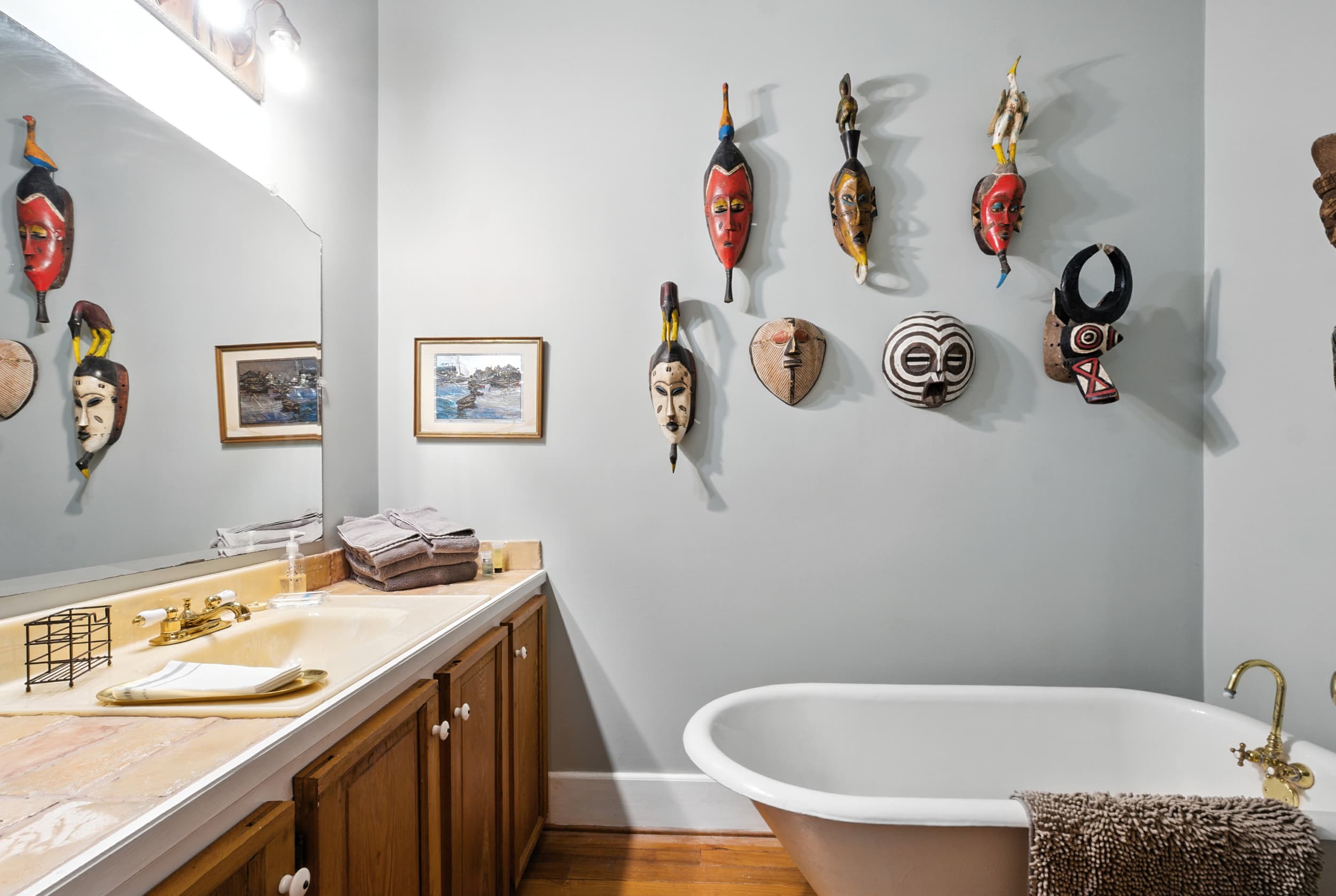 Eclectic bathroom featuring a collection of colorful tribal masks on the wall, a white freestanding tub, and a wooden vanity with a large mirror.