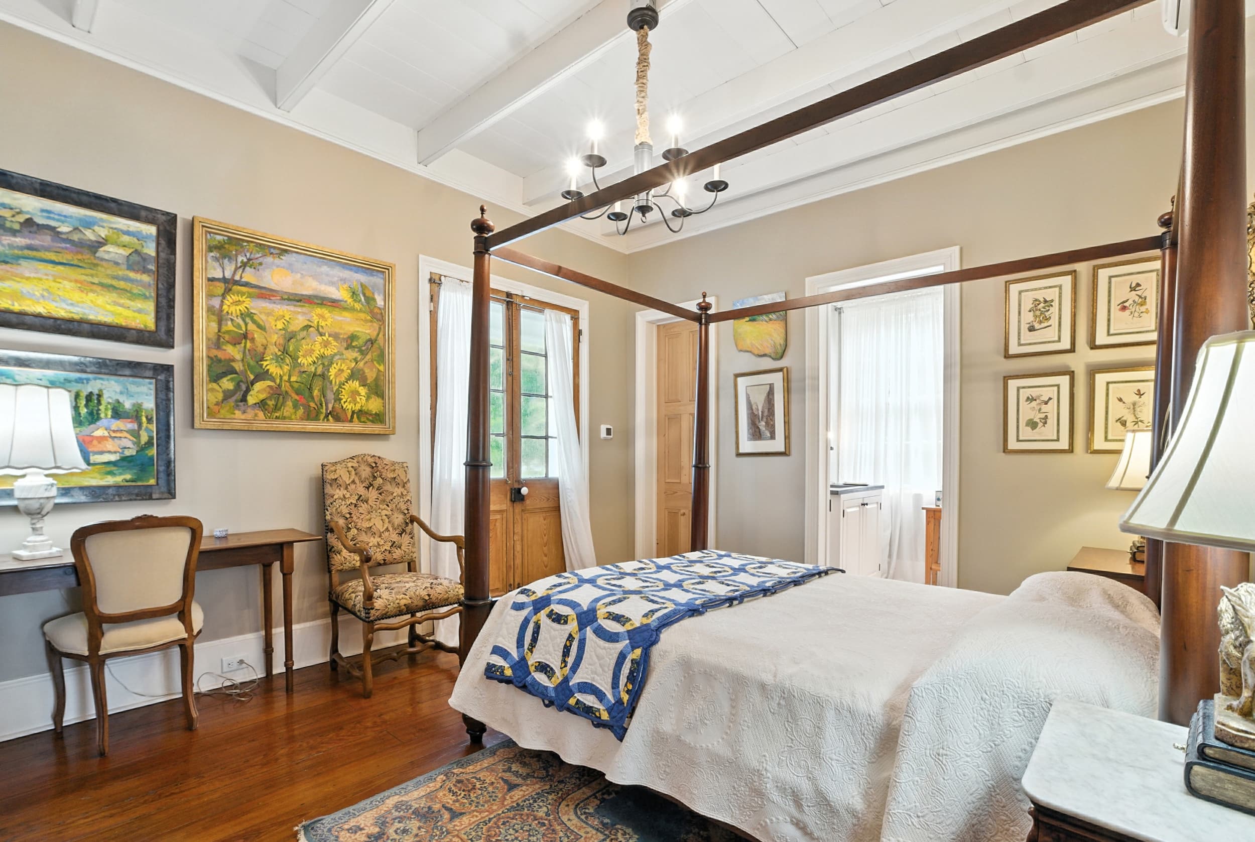 Elegant bedroom featuring a four-poster bed with a blue quilt, antique writing desk, and colorful landscape paintings on the walls.