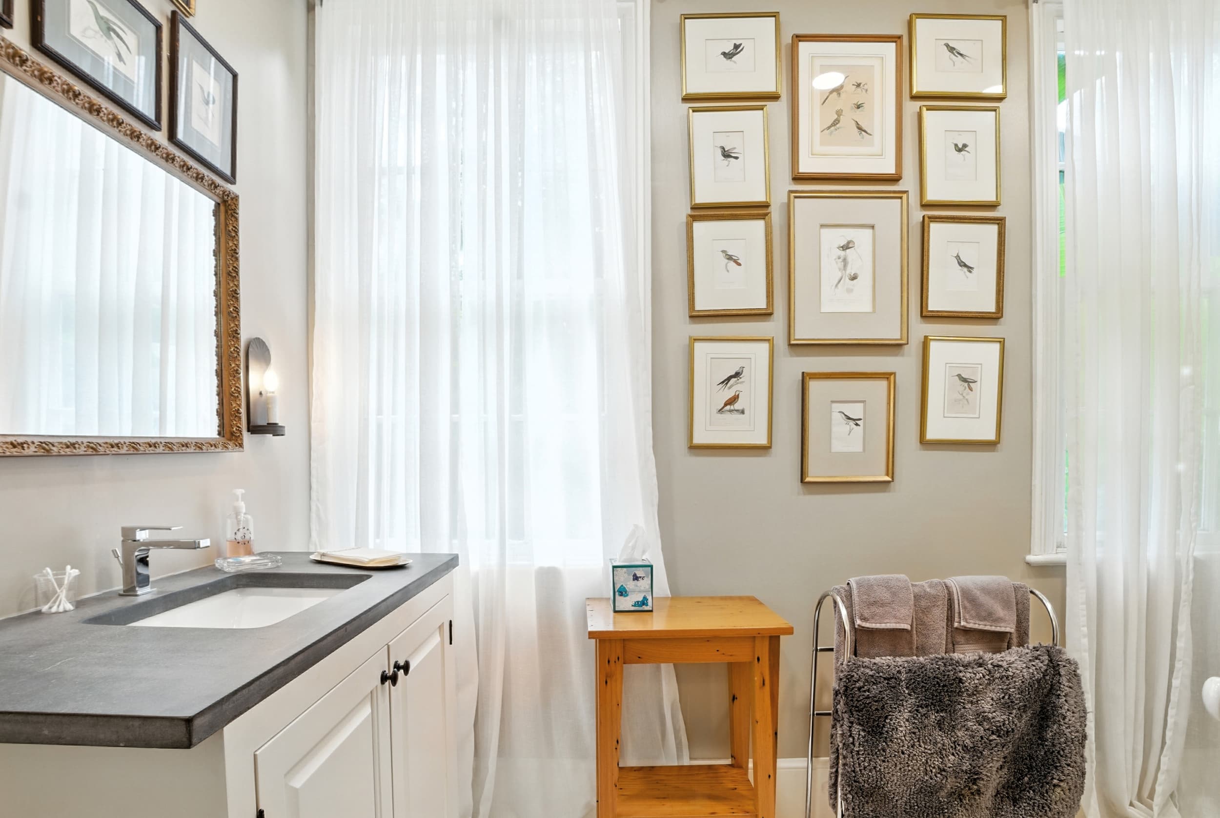 Bright bathroom featuring a vanity with a dark countertop, sheer white curtains, and a gallery wall of framed bird illustrations.