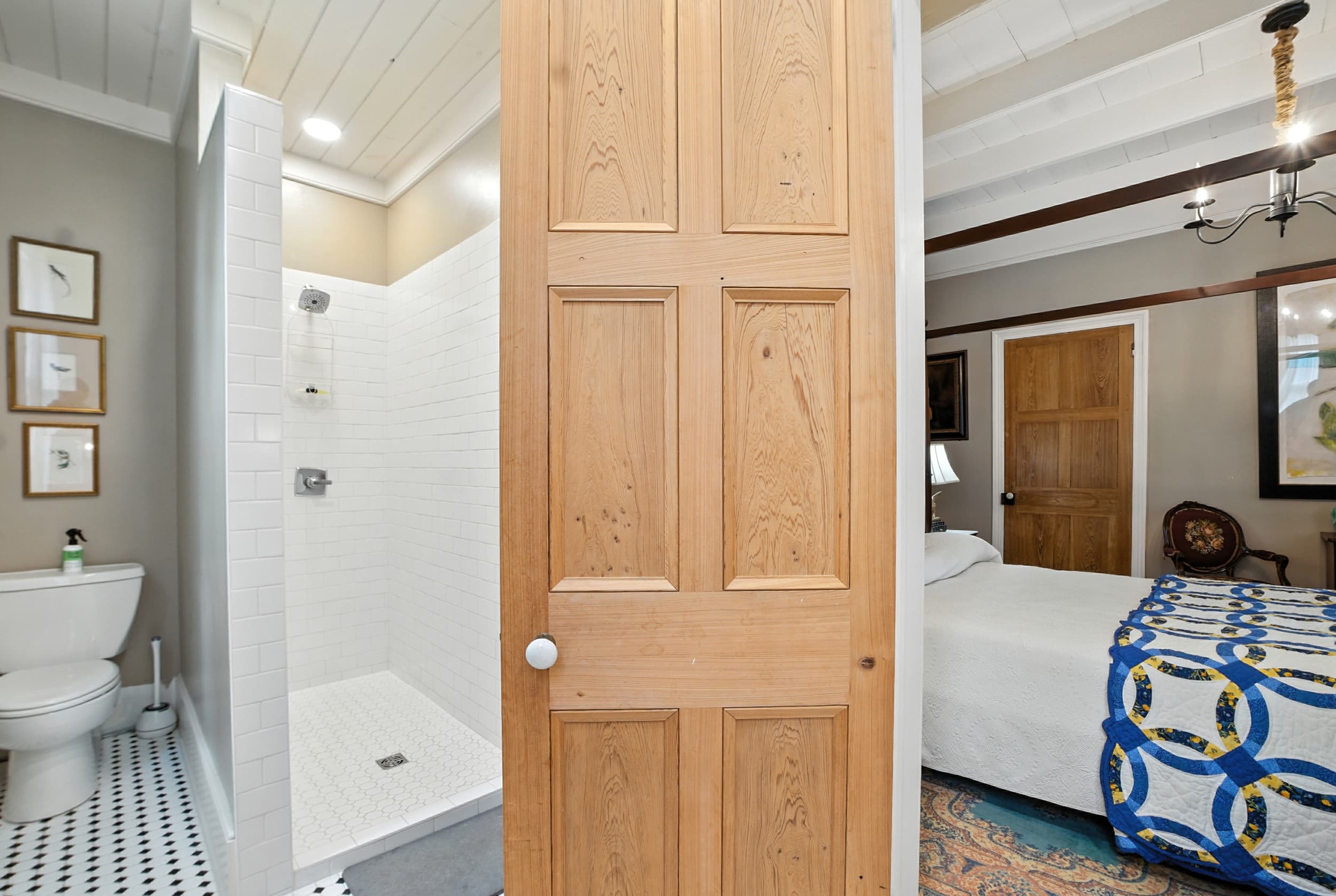 View of an ensuite bathroom with a walk-in tiled shower and black and white floors, adjacent to a bedroom with a blue and yellow quilt.