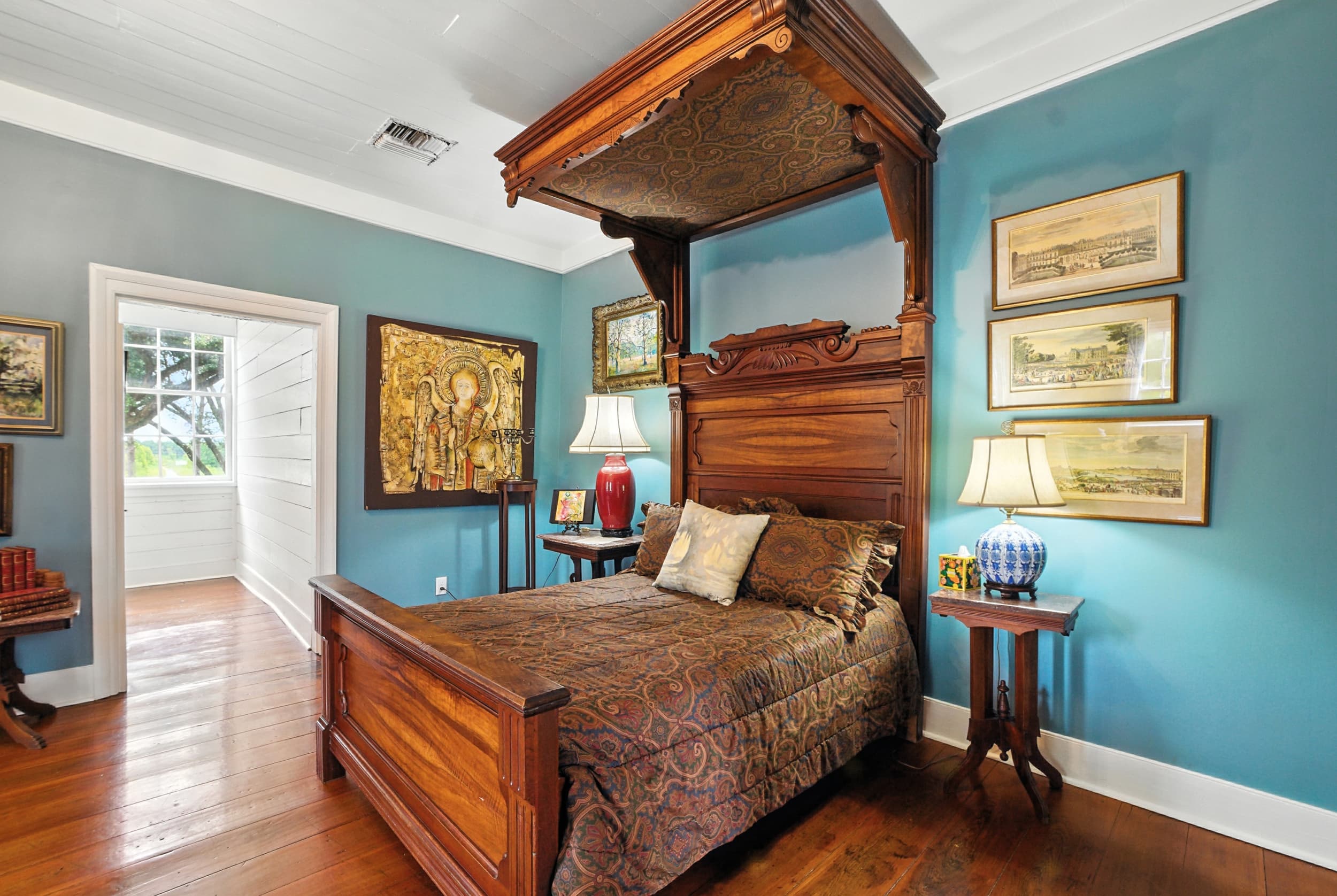 Teal bedroom featuring a grand antique wooden half-tester bed, hardwood floors, framed artwork, and a doorway leading to a sunlit porch.