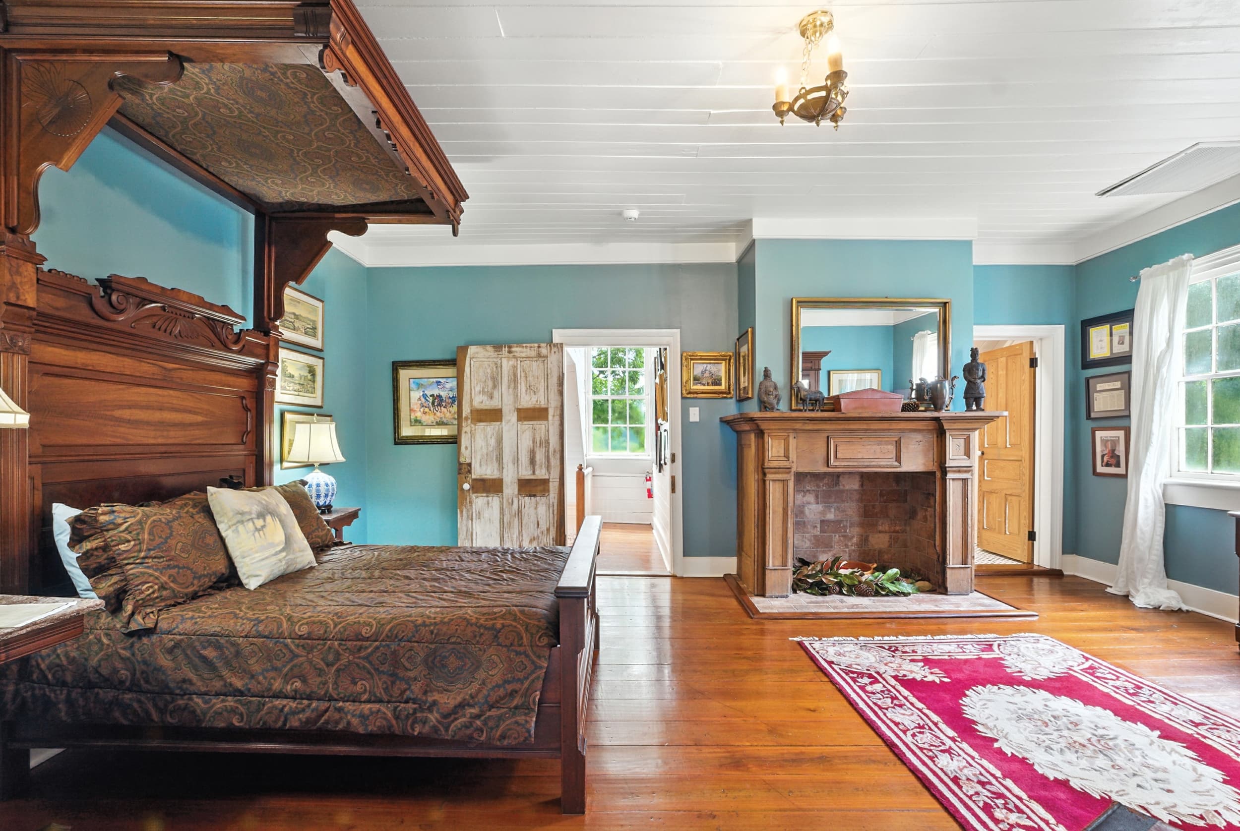 Spacious teal bedroom featuring a grand antique half-tester bed, a wooden fireplace mantel with a mirror, hardwood floors, and a red area rug.
