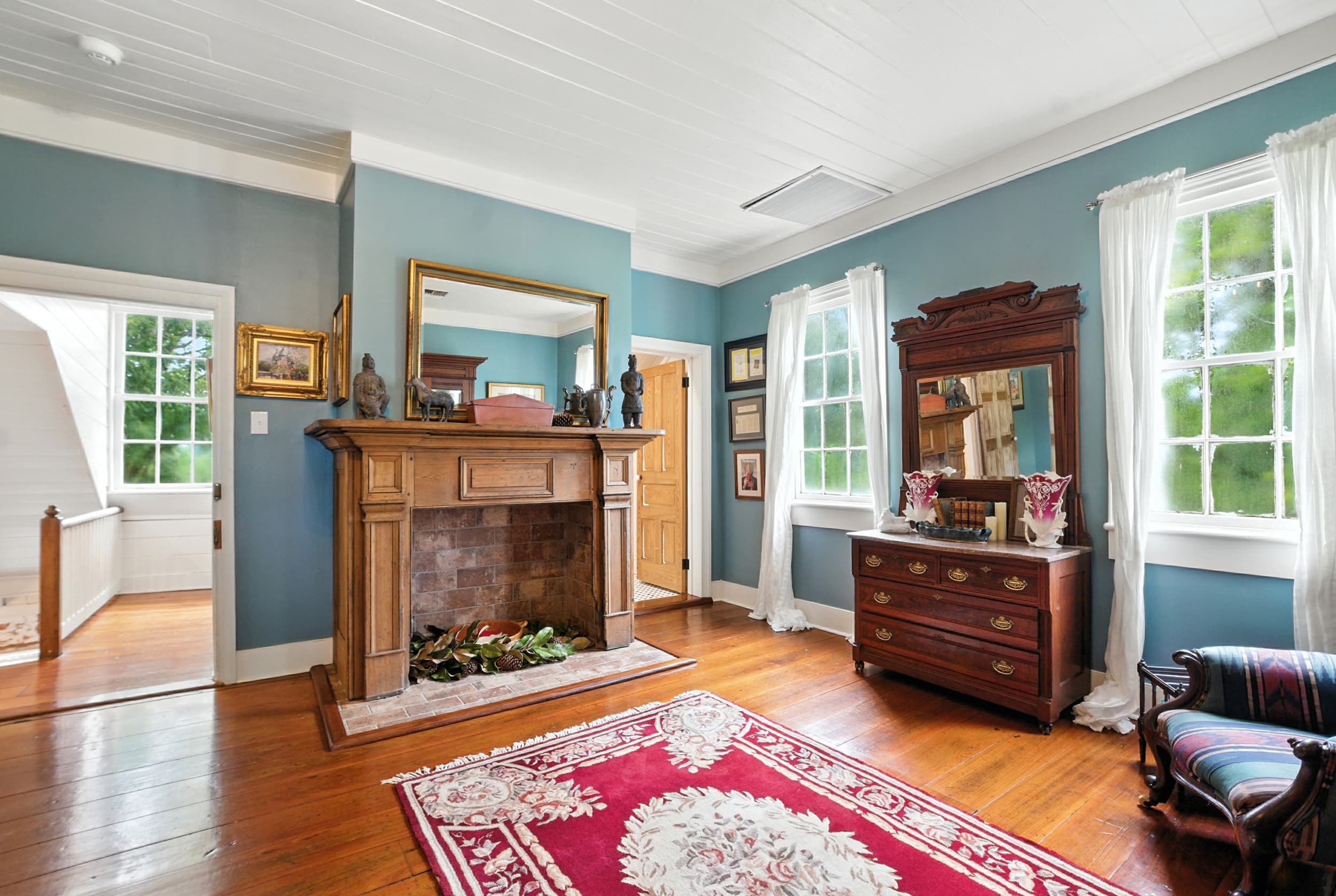 Spacious room with teal walls featuring a wooden fireplace mantel, antique dresser with mirror, hardwood floors, and a red patterned rug.