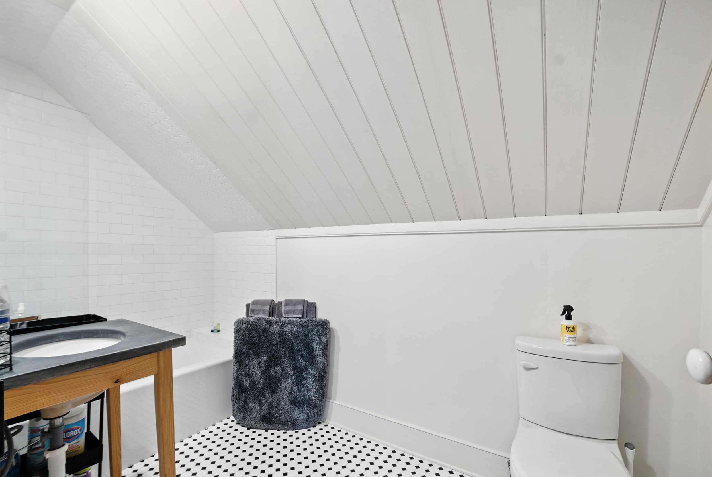 Bright bathroom featuring a sloped white plank ceiling, black and white geometric tile floors, a wooden vanity with a dark countertop, and a white toilet.