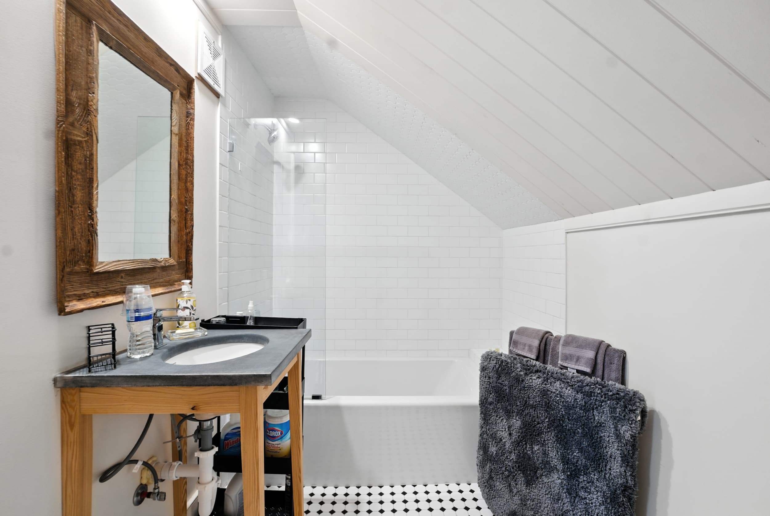 Bright bathroom featuring a sloped white plank ceiling, rustic wooden mirror, open vanity with a dark countertop, glass-enclosed tub, and geometric black and white tile floors.