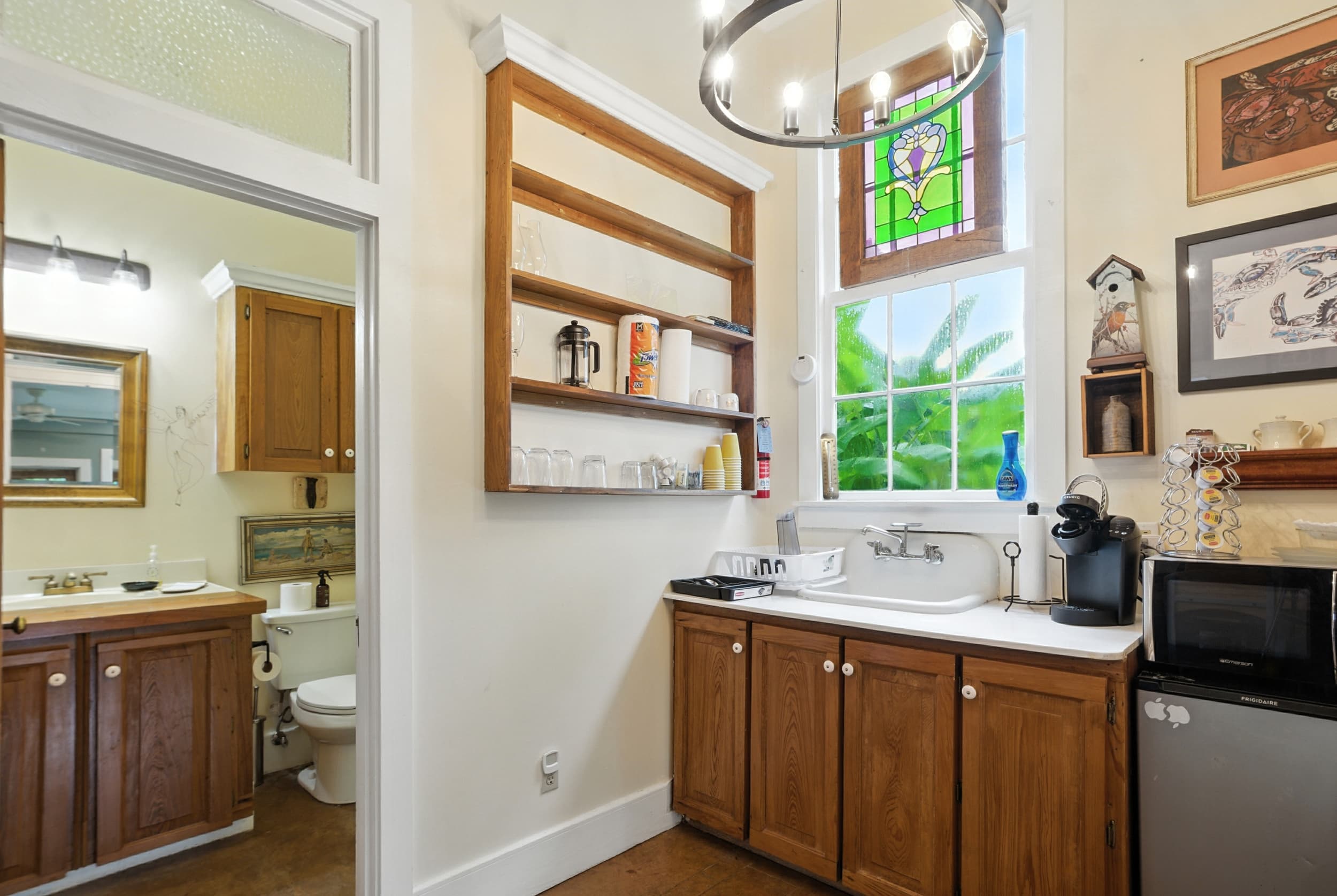 Cozy kitchenette featuring wooden cabinetry, open shelving, a stained glass window transom, small appliances, and a view into the adjacent bathroom.