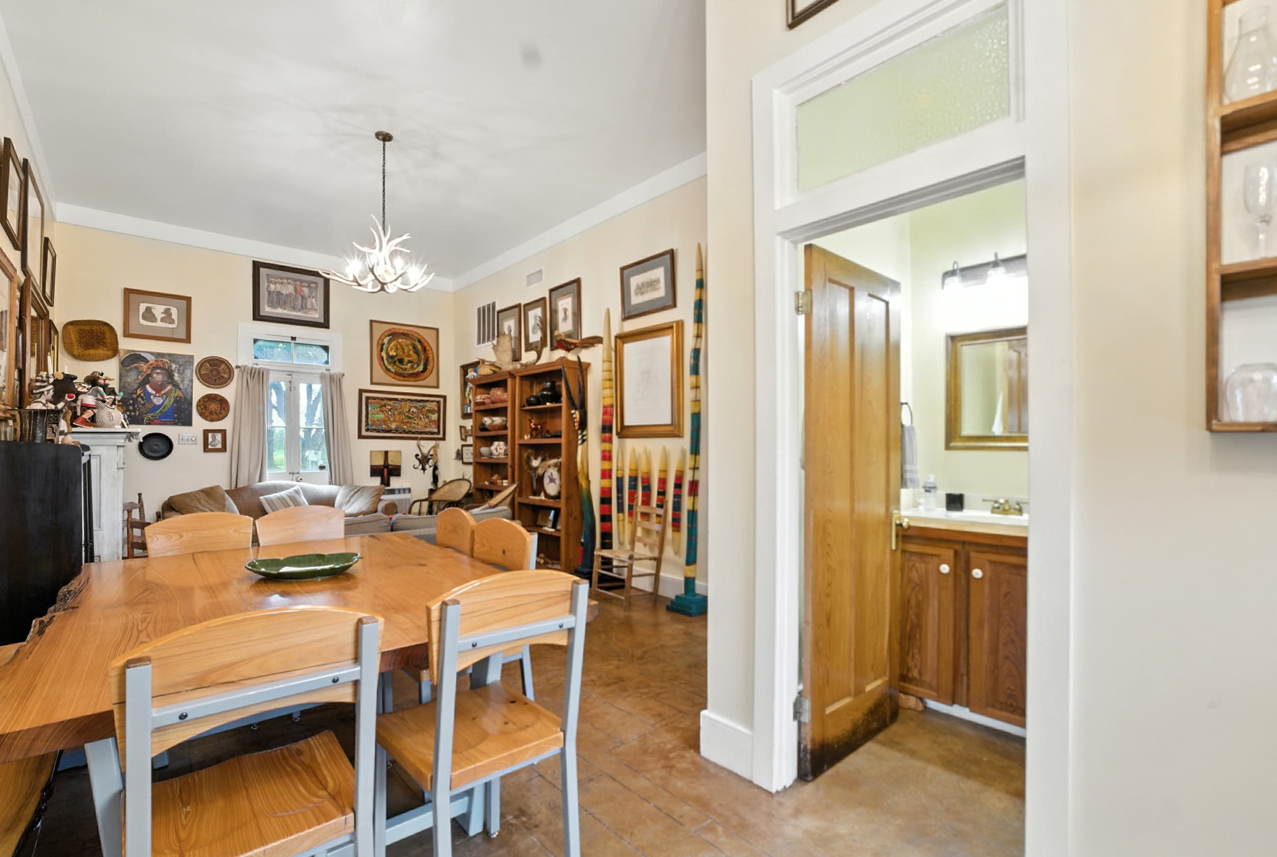 Eclectic living and dining area featuring a wooden table, a wall filled with framed art and artifacts, and a doorway leading to a bathroom.