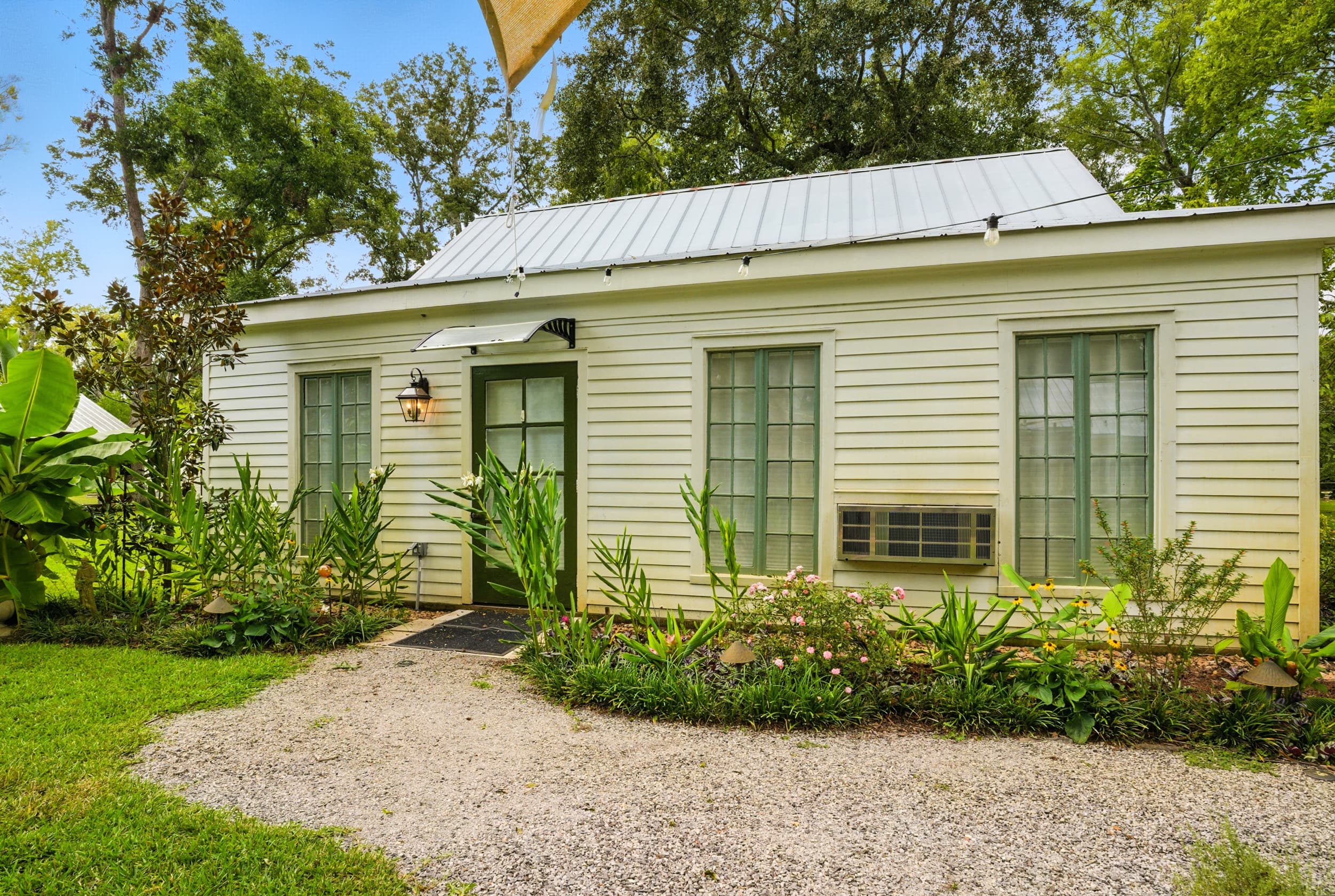 Exterior view of a charming white cottage with green trim, a silver metal roof, and a gravel path surrounded by lush greenery.