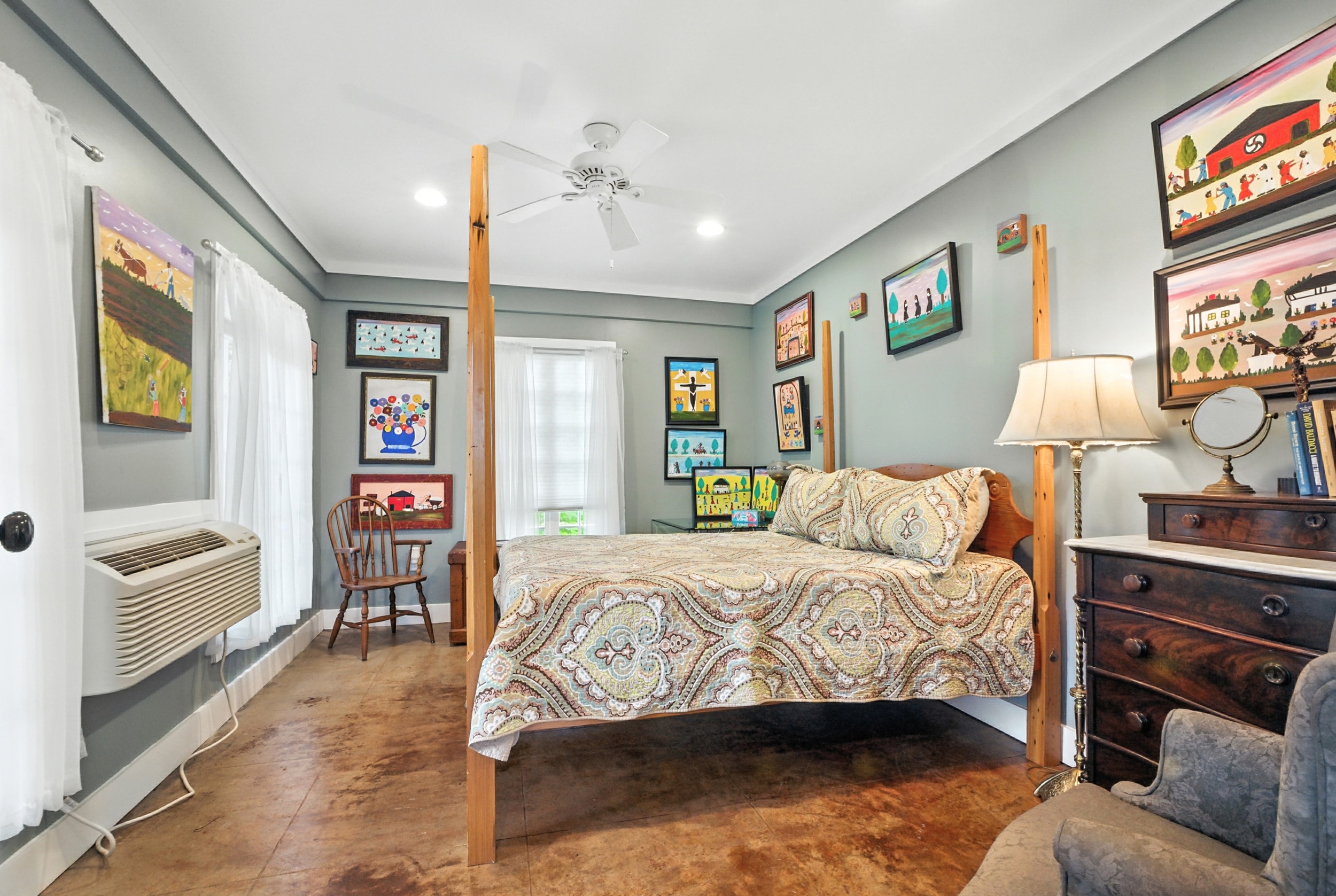 Eclectic bedroom featuring a wooden four-poster bed, a gallery wall of colorful folk art, an antique dresser, and rustic brown floors.