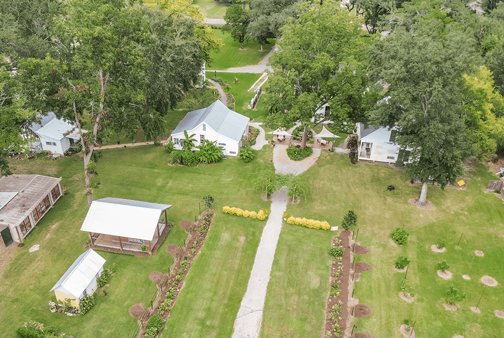 Aerial view of a lush green garden with pathways, surrounded by several quaint white buildings and large trees, evoking tranquility and charm.