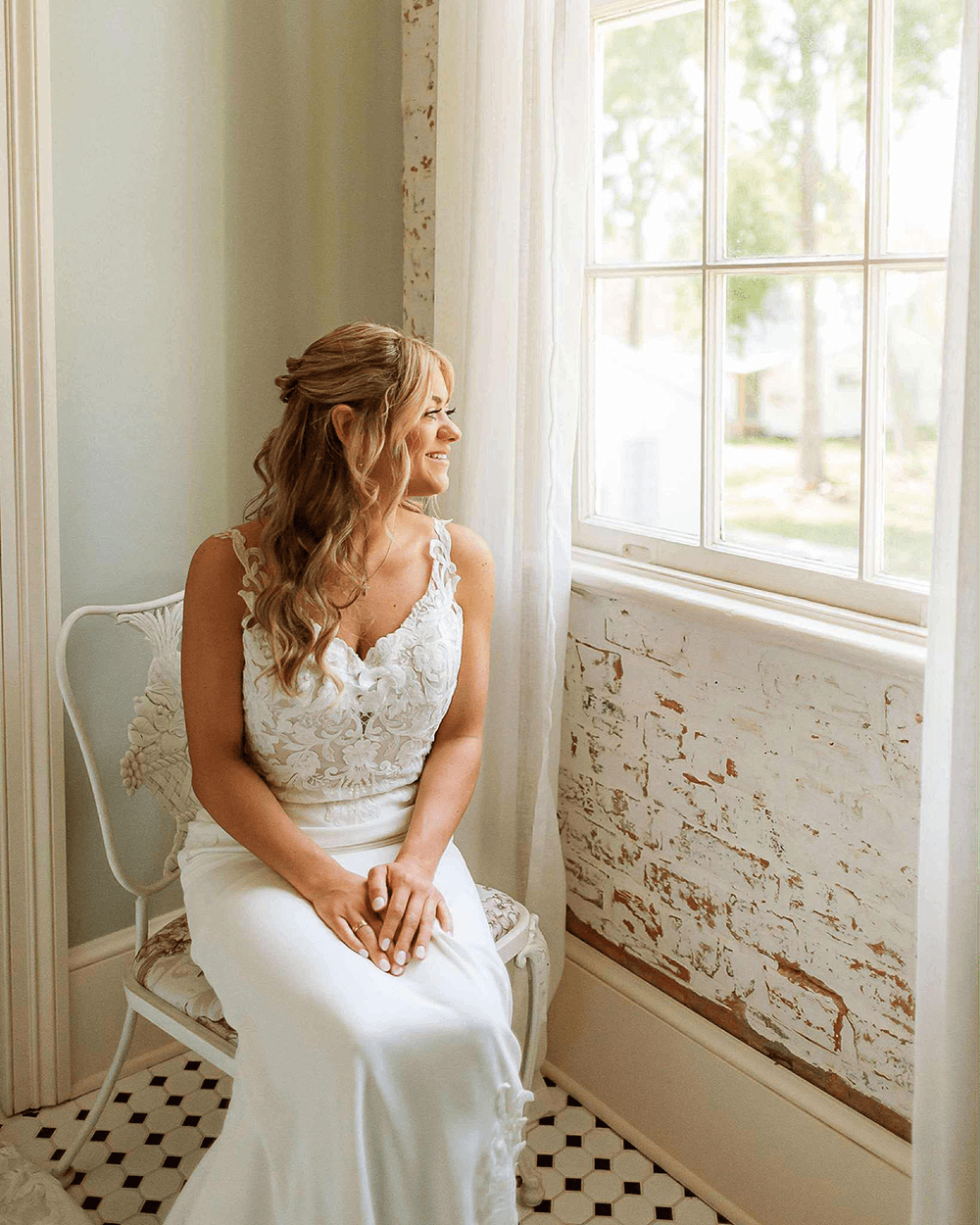 A woman in a white dress sits by a window, smiling as she gazes outside.