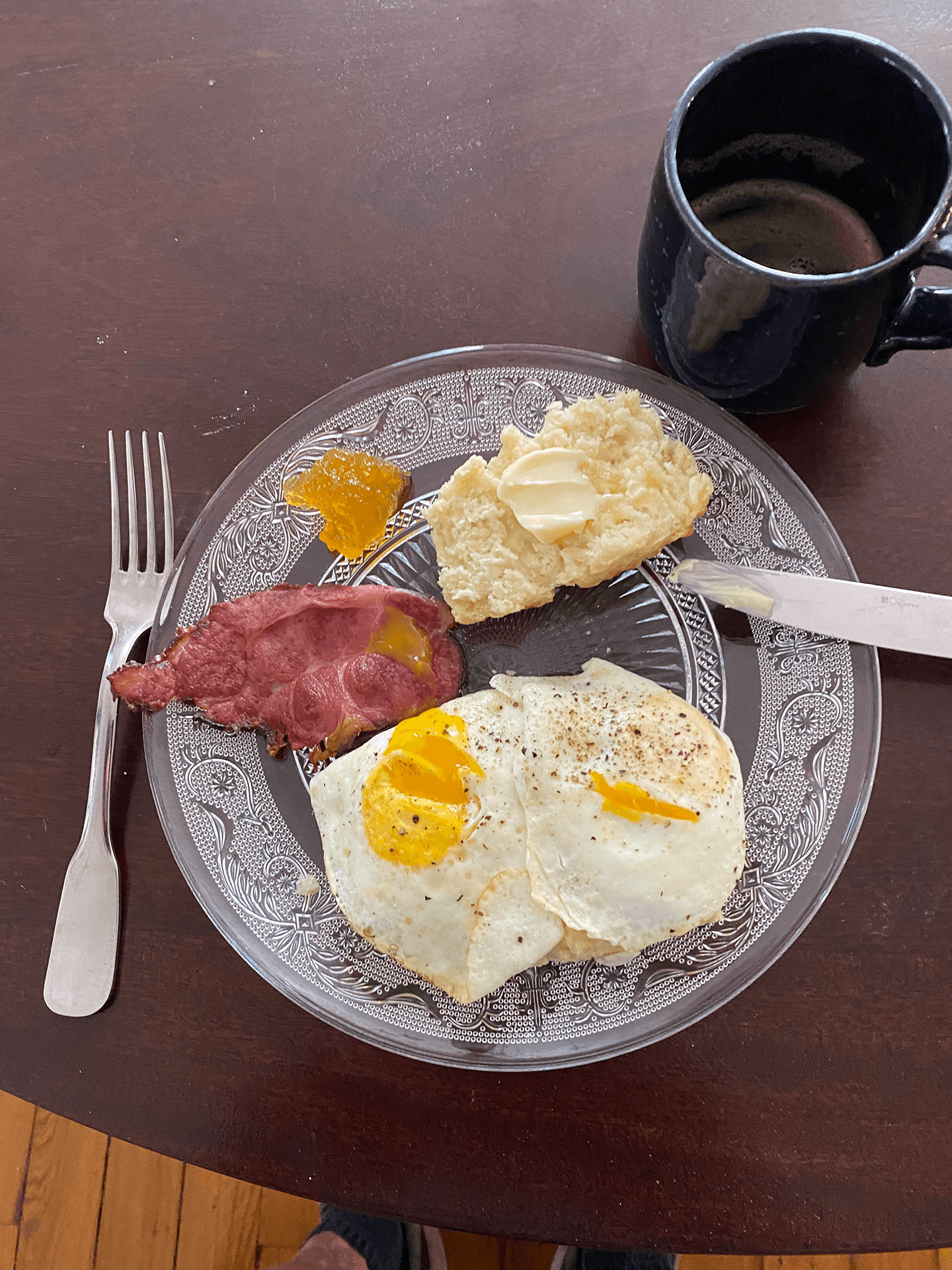 A plate featuring two fried eggs, a slice of bacon, a biscuit with butter, and a dollop of fruit preserves, alongside a coffee cup.