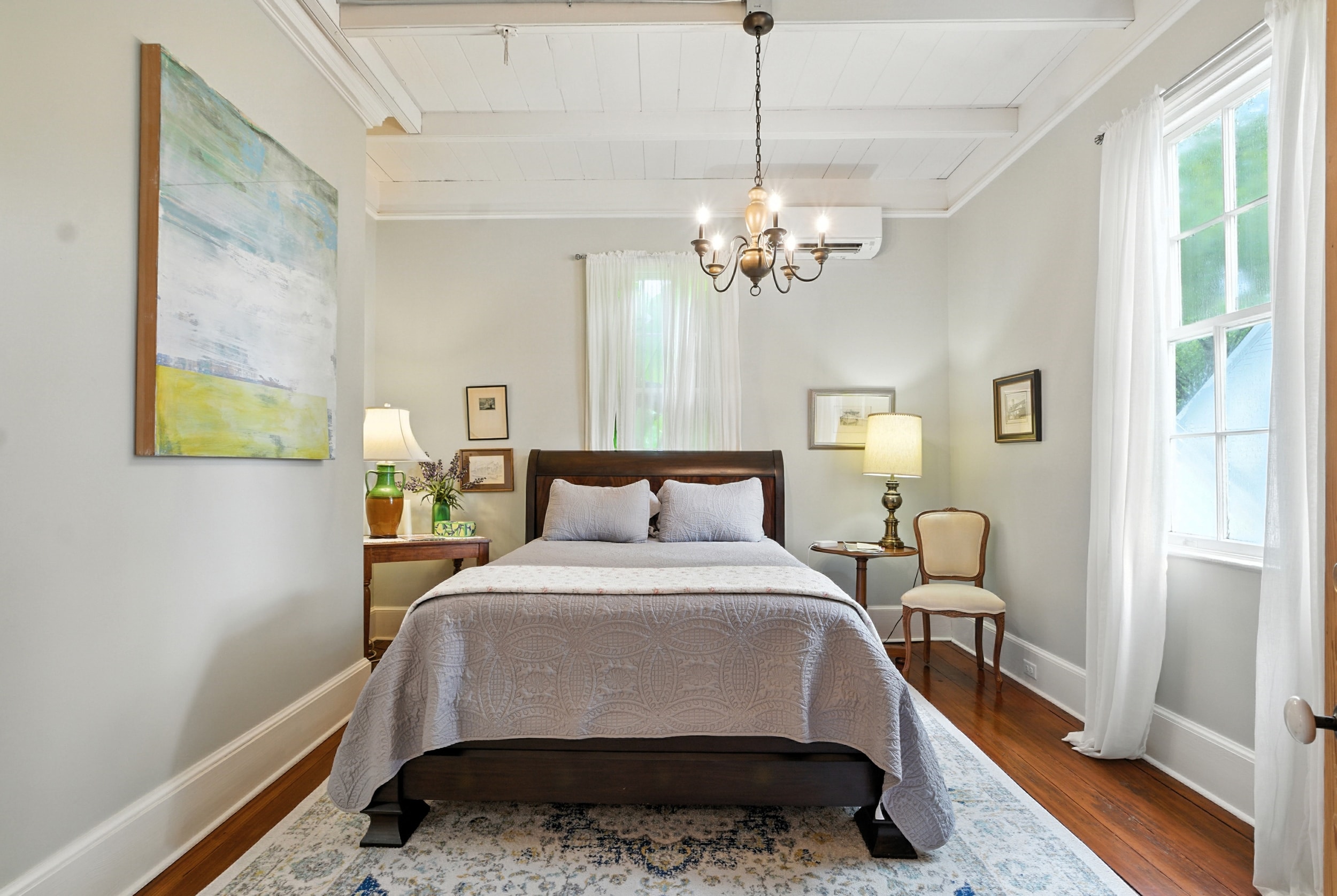 Bright bedroom featuring a dark wood sleigh bed, blue patterned rug, large abstract painting, and a chandelier hanging from white plank ceilings.