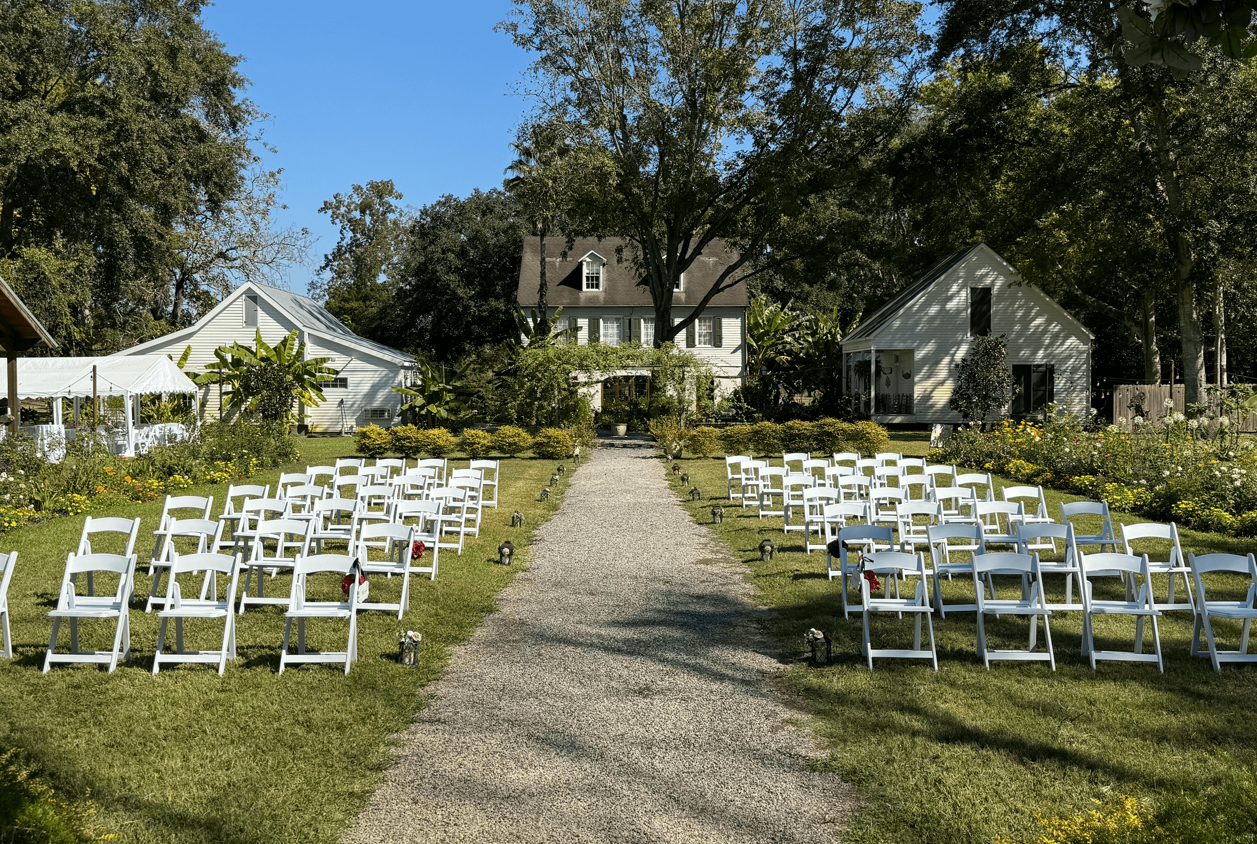 A pathway lined with white chairs leads to a decorative archway in a garden setting with two houses in the background.
