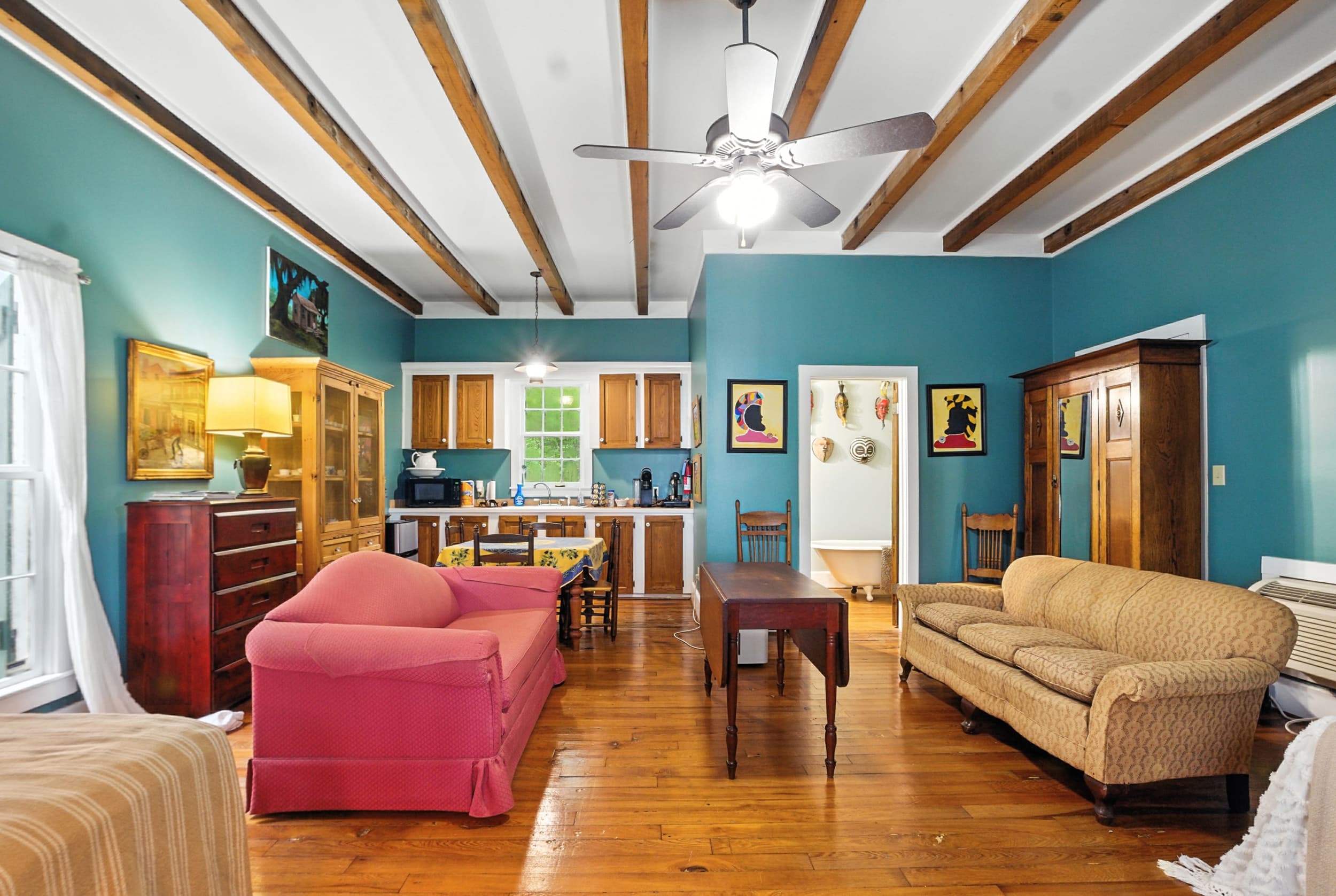 Spacious living area featuring deep teal walls, exposed wooden ceiling beams, a pink sofa, hardwood floors, and a kitchenette in the background.