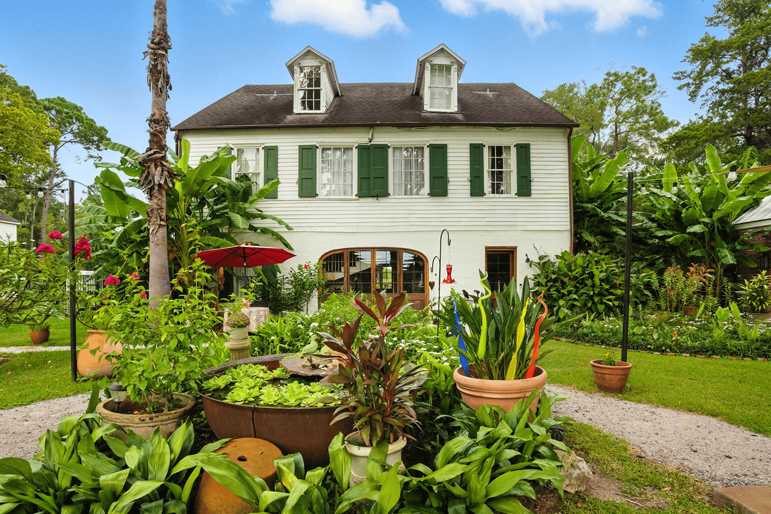 A charming two-story house with green shutters is set amid lush gardens. Potted plants, a red umbrella, and a bird feeder create a vibrant, serene scene.