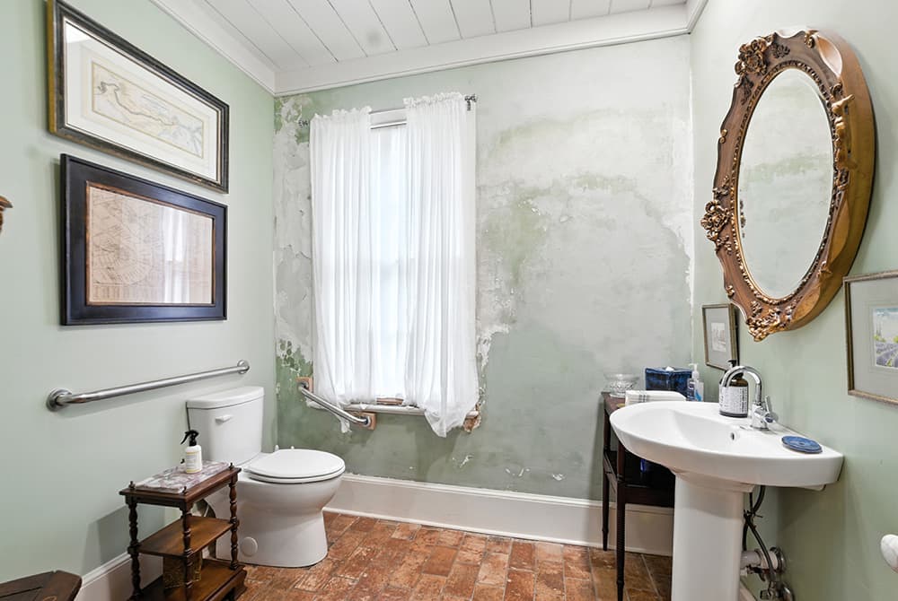 A small, partially finished bathroom with a toilet, sink, and large window featuring sheer curtains.