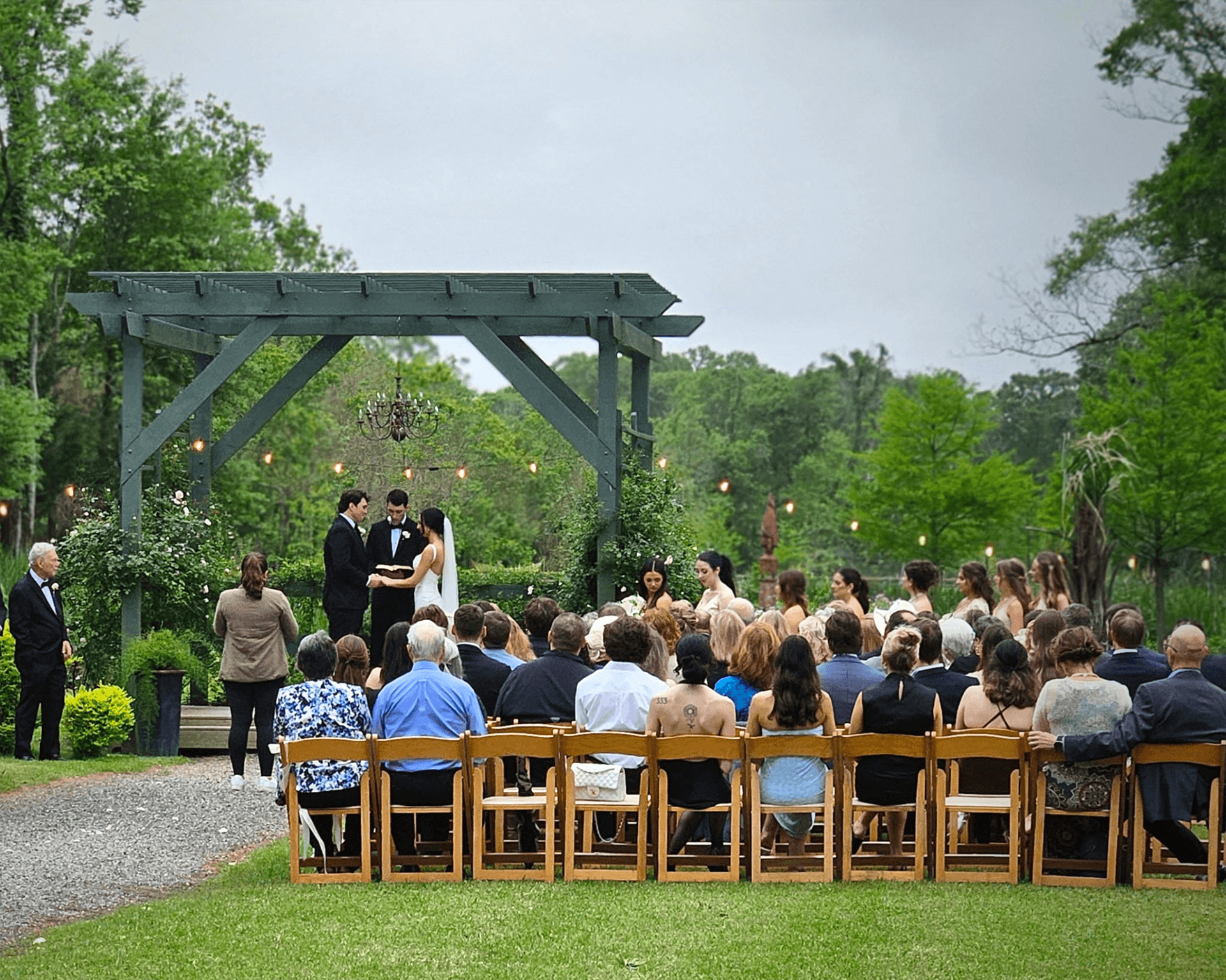 Outdoor wedding ceremony with a couple under a wooden arbor, surrounded by greenery. Guests seated on wooden chairs, creating a warm, intimate atmosphere.