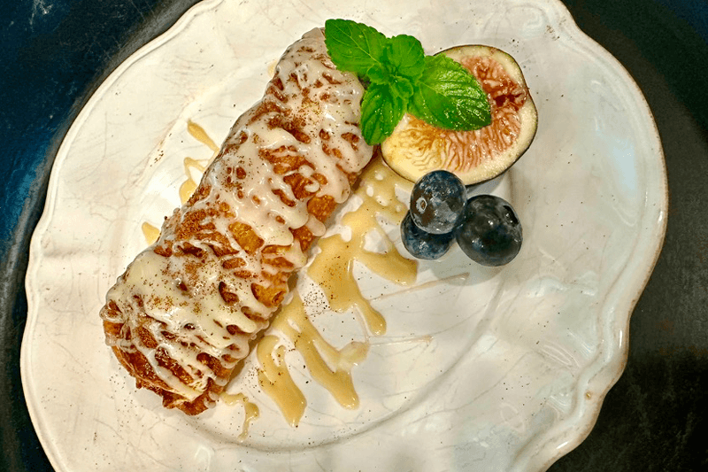 A crispy pastry drizzled with icing, garnished with a mint leaf and served with a fig and blueberries on a decorative plate.
