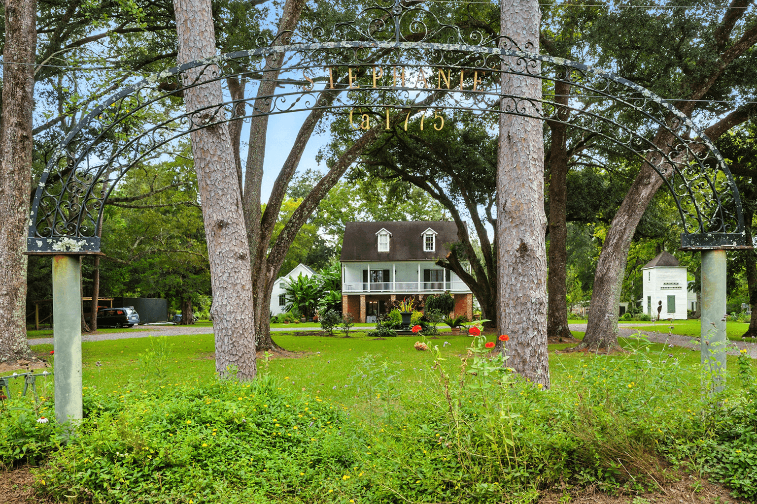 A charming two-story house with a dark roof and white balcony sits behind a wrought iron archway and lush garden, surrounded by tall trees.