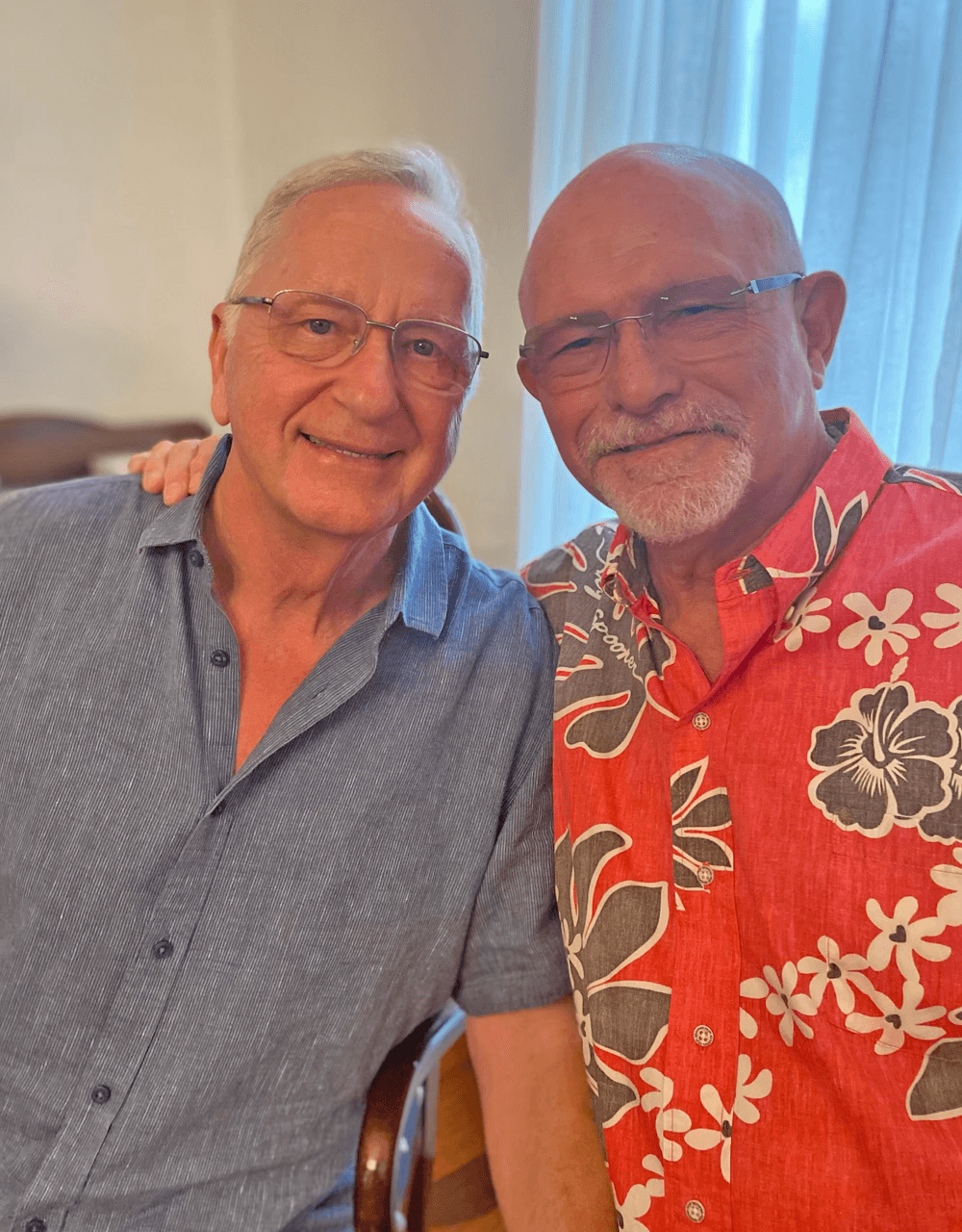 Two smiling men pose together, one wearing a blue shirt and the other in a red floral shirt.